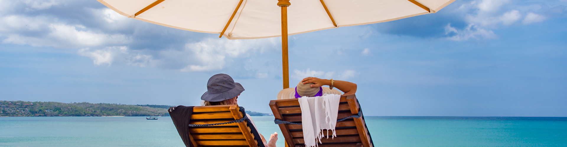 Twee personen zitten op een strandstoel met een parasol boven hen. Ze kijken over de zee.